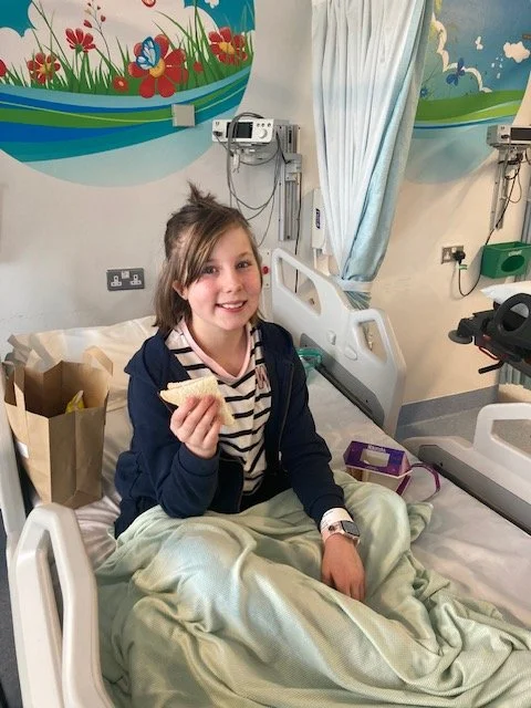A young girl sitting up in a hospital bed, smiling and holding a sandwich, with medical equipment and colorful wall art in the background.