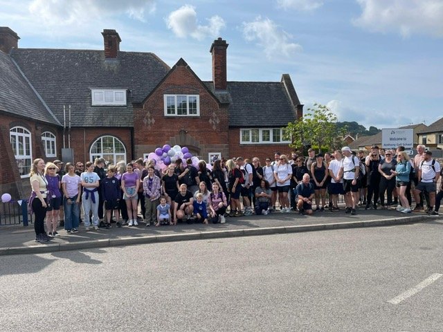 Group of children and adults gathered outside a brick building with balloons and a sign in the background.