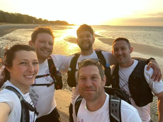 Group of five friends taking a selfie on the beach during sunset, all wearing matching white shirts and backpacks.