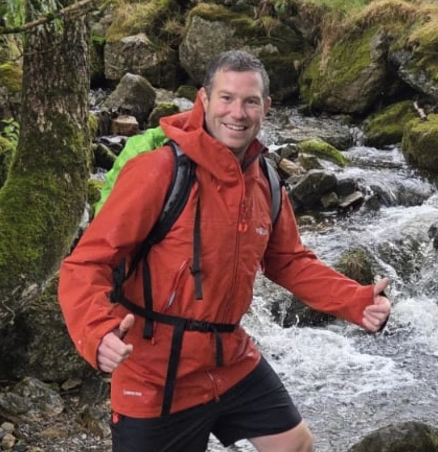 A man in a red rain jacket and black shorts smiles and gives a thumbs-up while standing in a rocky stream in a forest.
