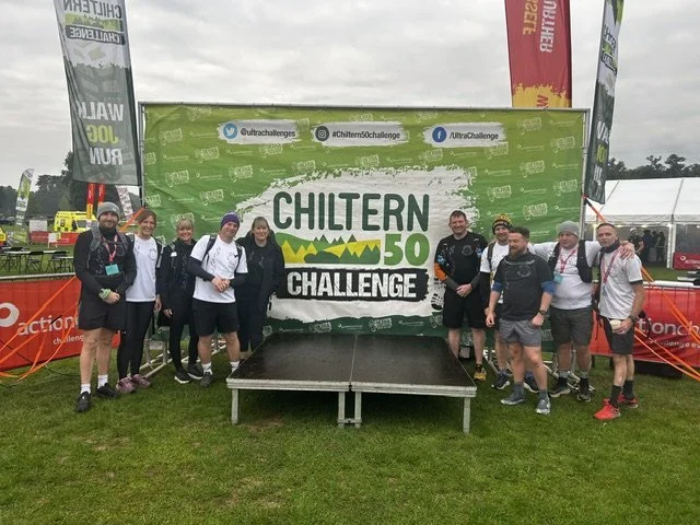 Group of people standing on a stage in front of a green banner that says 'Chiltern 50 Challenge' with social media handles at the top. The event appears to be outdoors on a cloudy day.