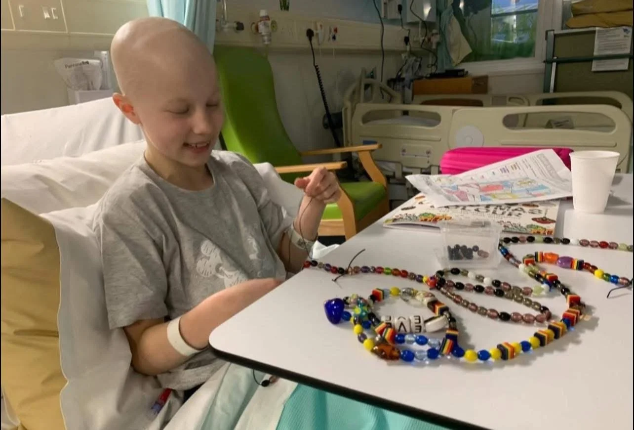 A young child with a shaved head in a hospital bed, wearing a hospital wristband, smiling at a table with colorful beaded necklaces and an open coloring book.