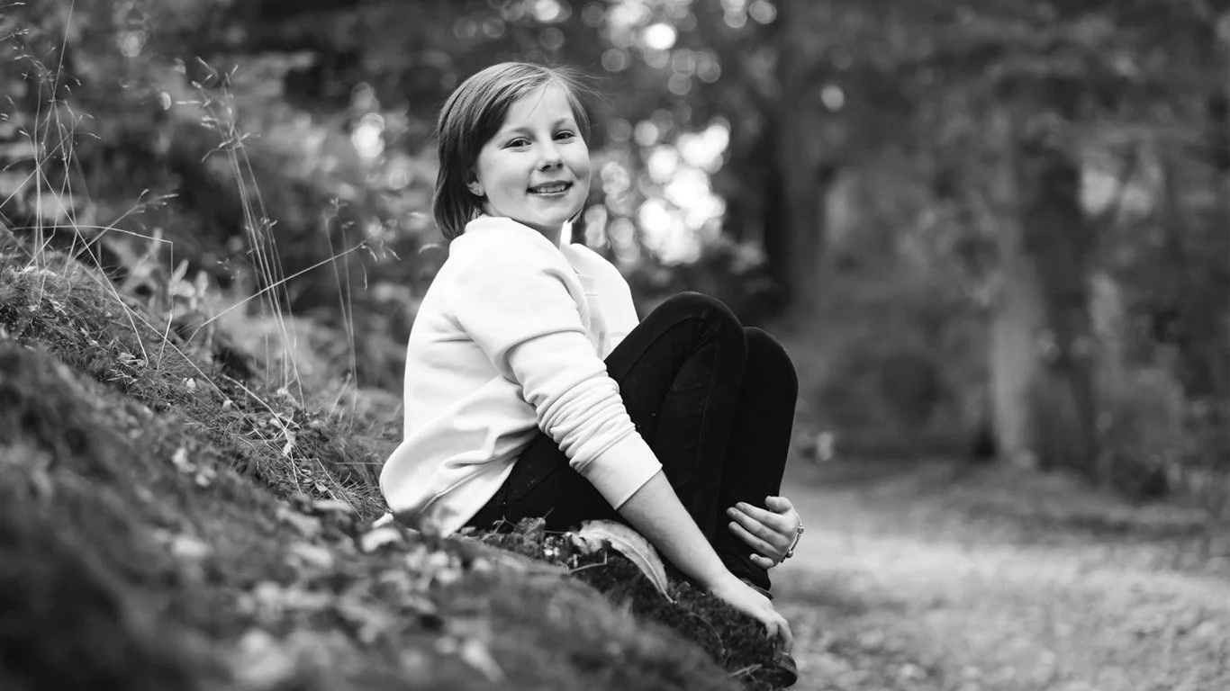 A young girl sitting on a grassy hill outdoors, smiling at the camera, with trees in the background.