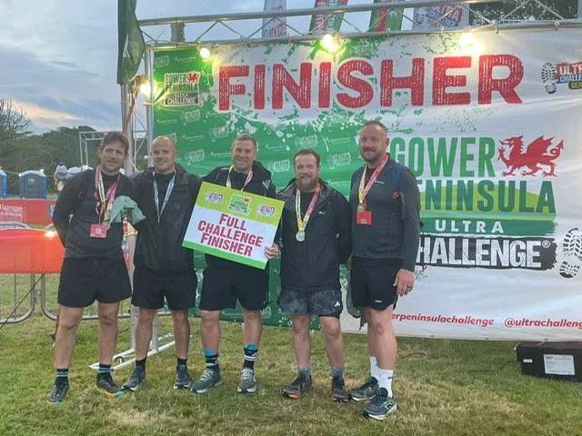 Group of five men standing in front of a banner that says "FINISHER POWER PENINSULA ULTRA CHALLENGE," each wearing medals, with one holding a sign that reads "FULL CHALLENGE FINISHER." They are outdoors at a race event.