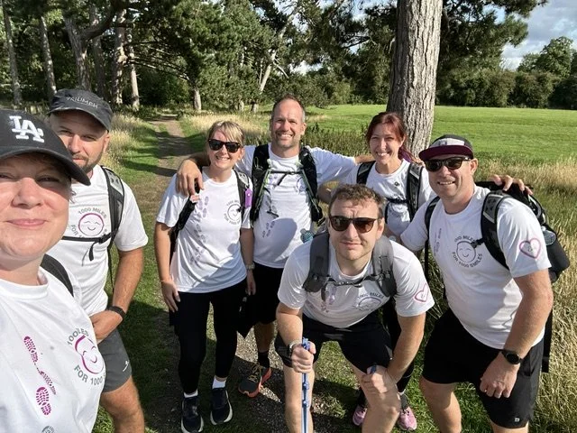 Group of seven people posing outdoors on a trail, smiling, with trees and grass in the background. They are wearing white T-shirts with a logo, backpacks, and sunglasses, indicating they are on a hike or outdoor activity.