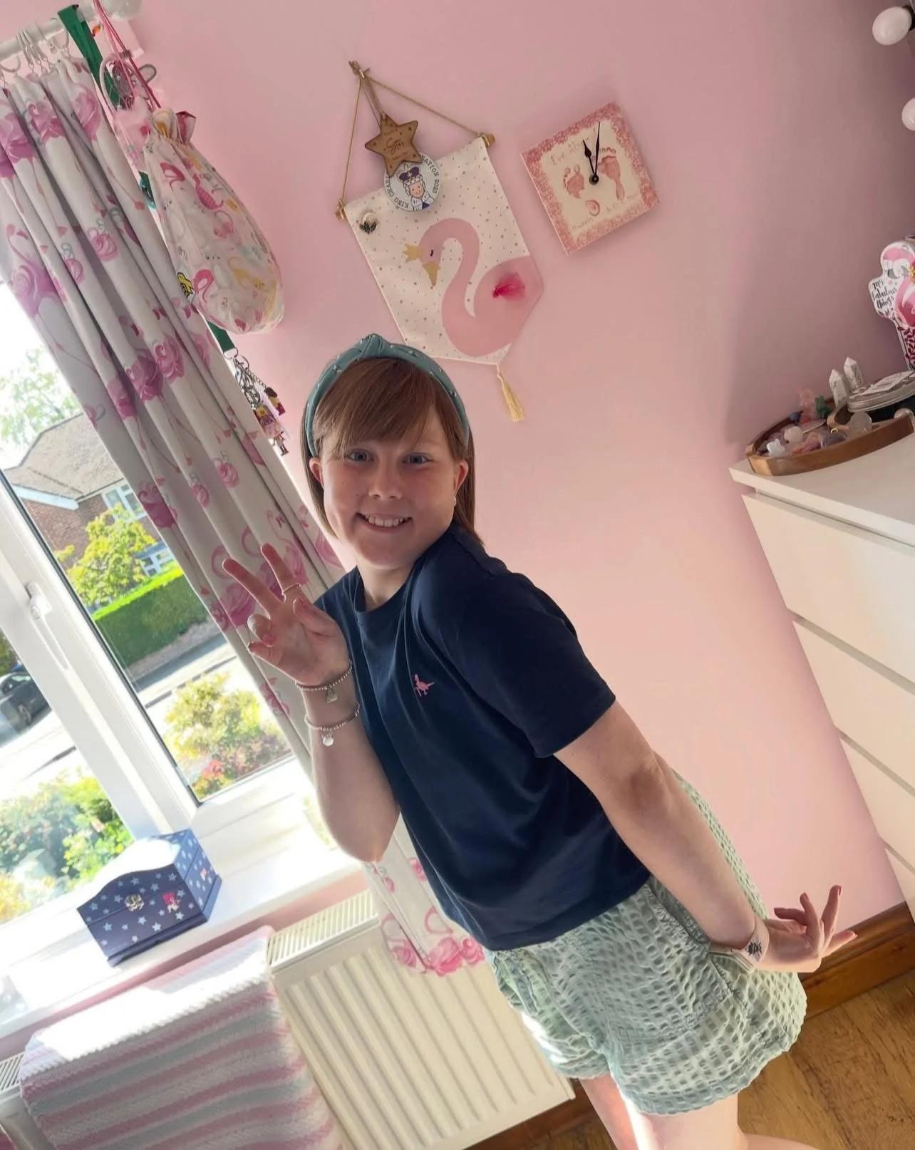 A young girl smiling and making a peace sign with her fingers inside a pink-walled room. The room has a window with pink curtains featuring flamingo designs, a swan-themed wall decor, and a dresser with a tray of small items.