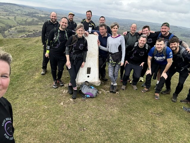 Group of people hiking on a grassy hilltop with a large stone marker, scenic landscape in the background.
