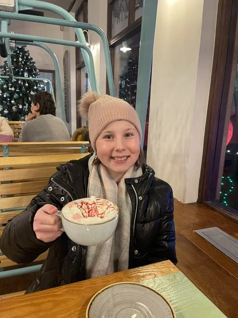 A young girl with a pink beanie, black jacket, and gray scarf sits at a wooden table in a restaurant, holding a mug of hot chocolate topped with whipped cream and red sprinkles, smiling at the camera. In the background, there is a decorated Christmas tree and other people dining.