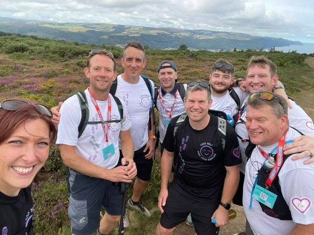 Group of eight people hiking outdoors in a landscape with purple heather and hills in the background, all smiling at the camera.