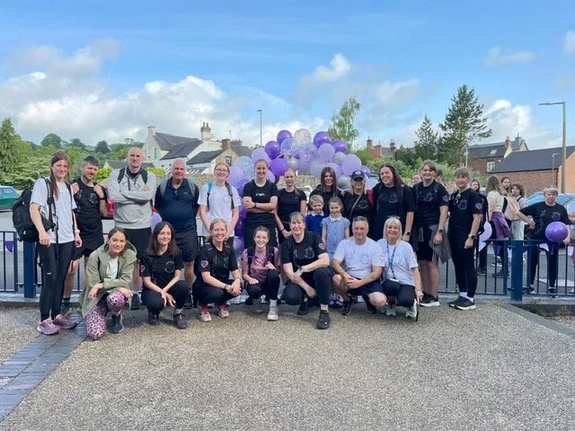 Group of students and adults posing outdoors with purple balloons in the background.