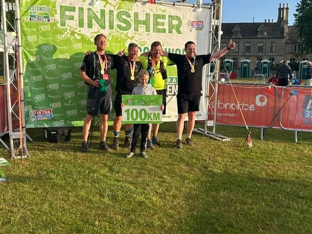 Four men and one young boy standing in front of a green backdrop at a race event, all wearing medals. The boy is holding a large sign that says '100 KM'. The men are celebrating with one giving a thumbs-up. The background shows a grassy area, a build