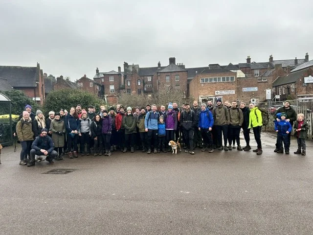 Group of people outdoors in a cloudy weather, standing on a paved area in front of houses, with children and a dog, dressed in outdoor clothing.