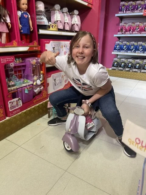 Girl smiling and squatting on a small pink and white toy scooter in a toy store, with shelves of toys behind her.