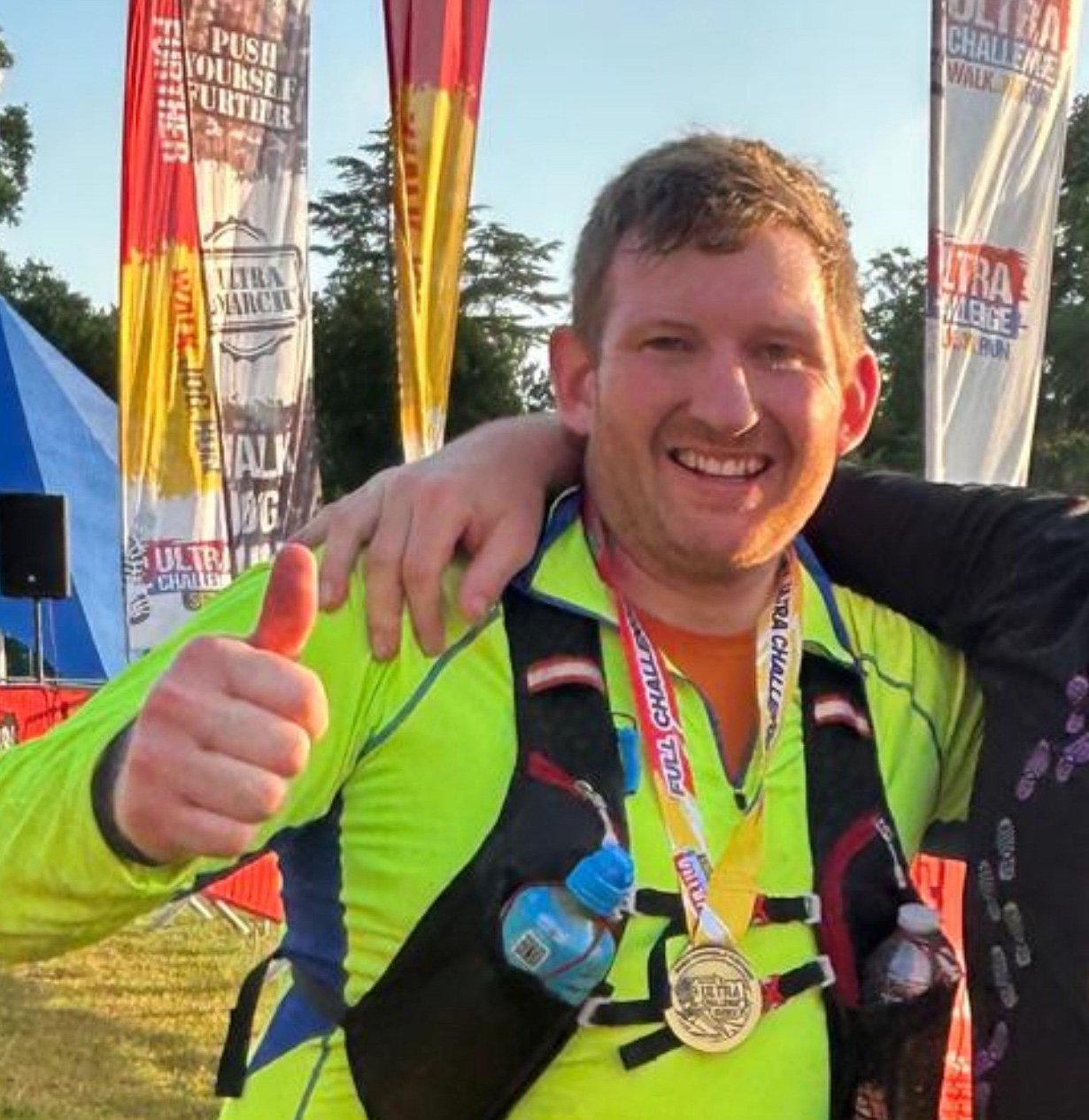 A smiling man wearing a neon green running shirt with a medal around his neck, giving a thumbs-up. He has a water bottle attached to his vest and an arm around someone next to him. In the background, there are colorful banners and tents, indicating an outdoor sporting event.