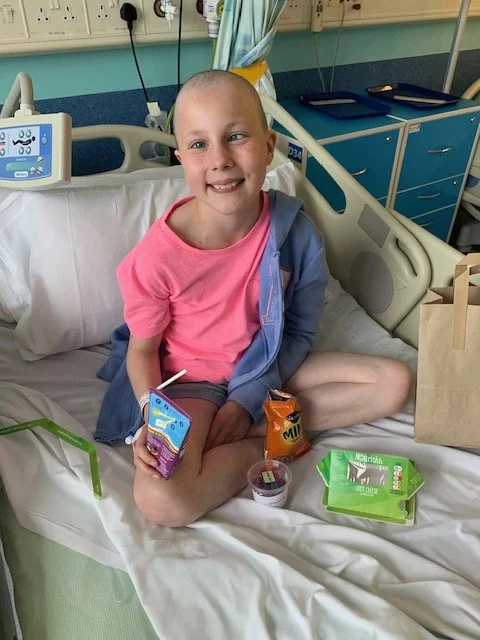 A young girl with a shaved head sitting on a hospital bed, smiling, holding a snack pack, surrounded by snacks and a paper bag, in a hospital room.