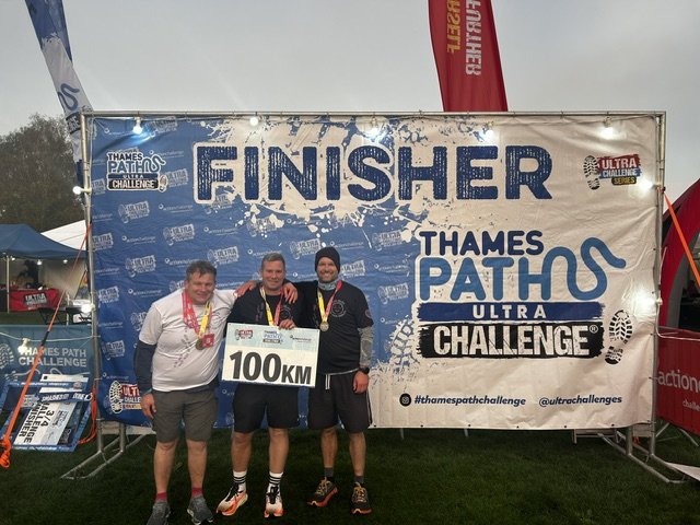 Three men standing in front of a large banner that says "FINISHER THAMES PATH ULTRA CHALLENGE" and holding a sign that says "100KM." They are smiling, wearing medals, and dressed in athletic gear, celebrating completing the ultra challenge.