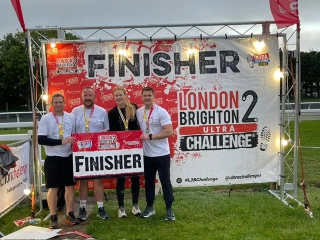 Four runners holding a 'Finisher' sign after completing the London Brighton 2 Ultra Challenge, standing in front of a large finish banner with event details.