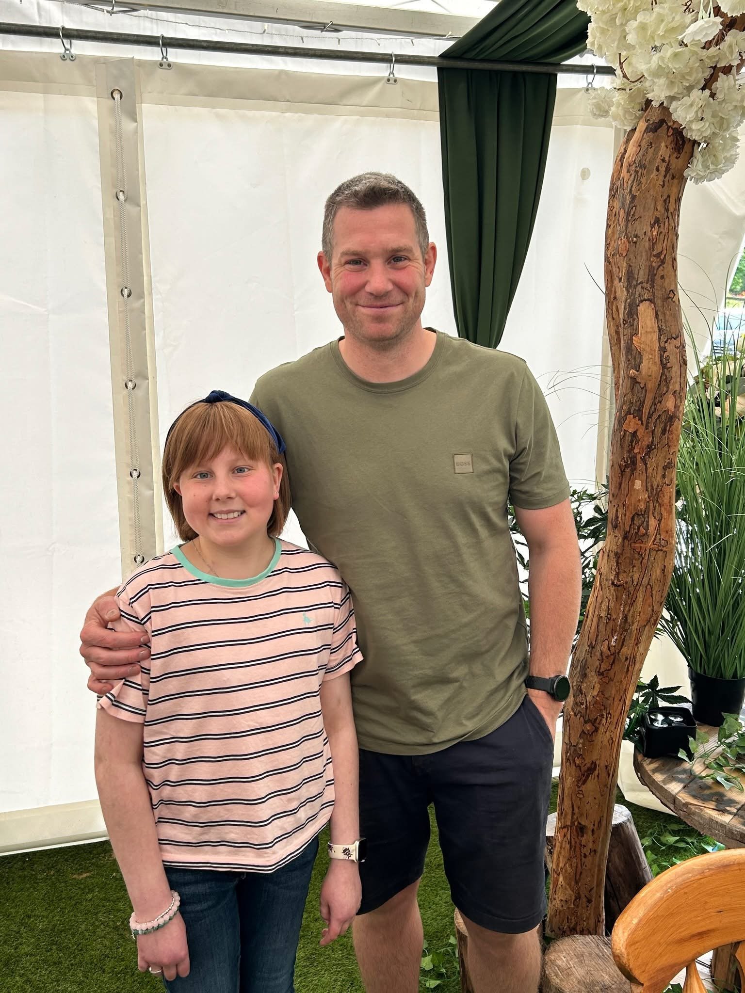 A man and a girl smiling at the camera inside a tent or greenhouse, with trees and plants around them, and a large decorative tree branch with white flowers to the right.