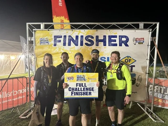 Group of five people celebrating after completing a race, standing under a finish line banner that reads 'FINISHER'. The person in the center holds a sign that says 'FULL CHALLENGE FINISHER.' It appears to be at night with event tents and banners aro