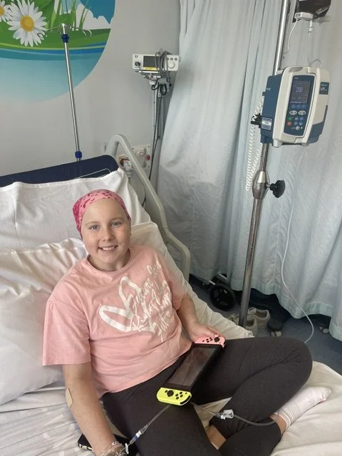 Young girl in hospital bed smiling, wearing a pink headscarf and pink shirt, with medical equipment nearby.