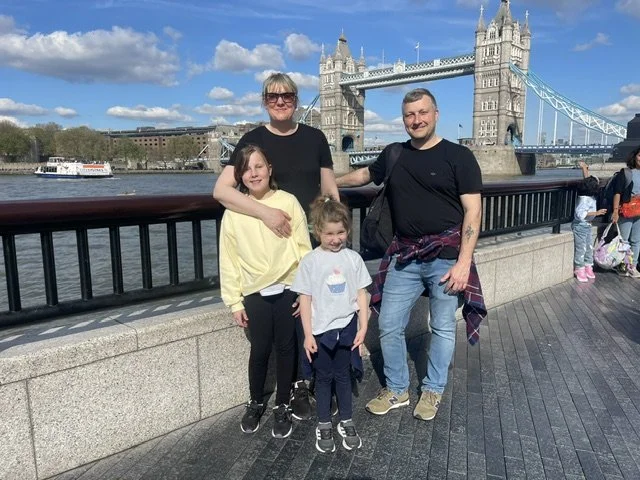 A family of four smiling for a photo near the Tower Bridge in London with the River Thames in the background, sunny weather, and other tourists nearby.