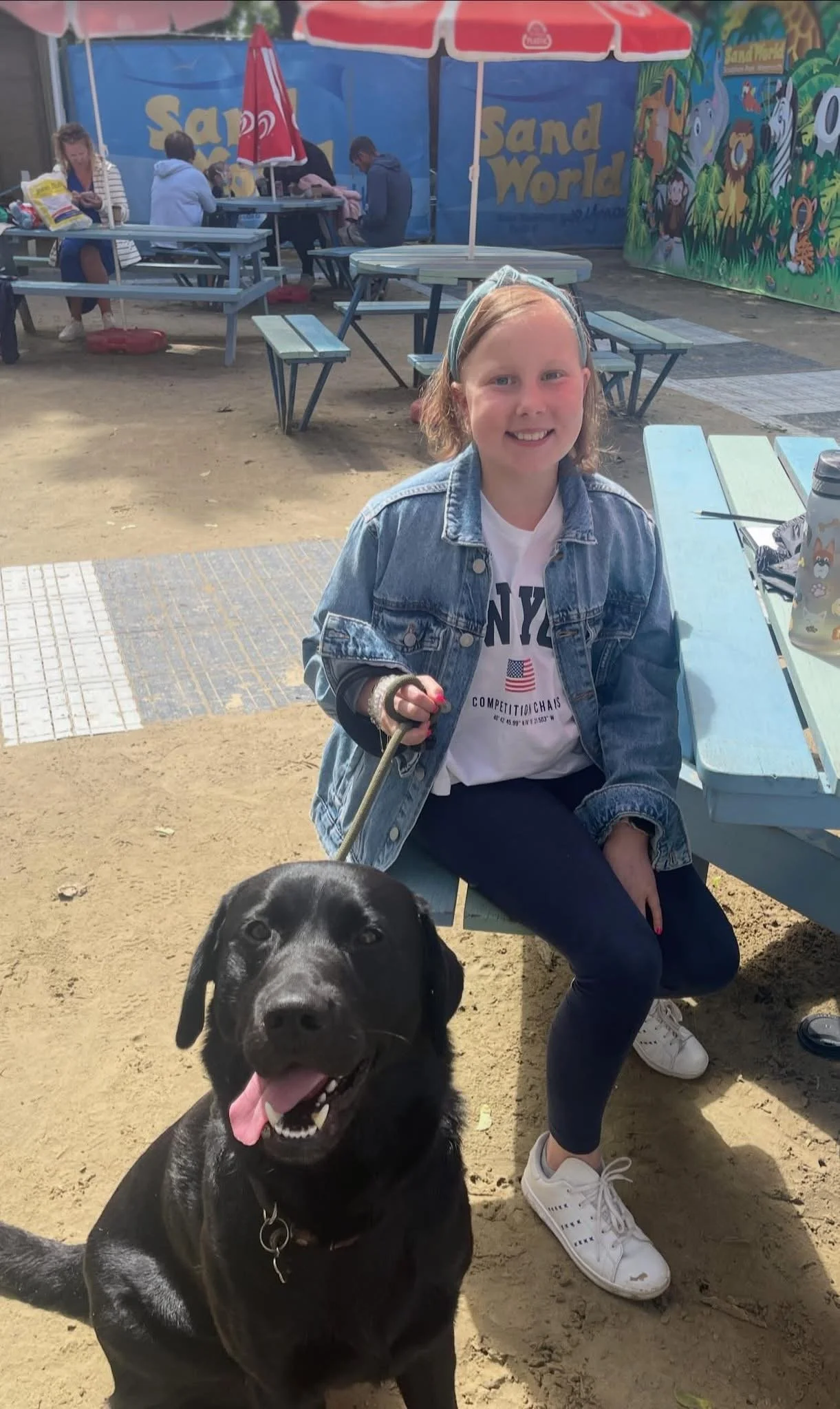 A young girl with glasses, wearing a denim jacket and white sneakers, is sitting at a picnic table outdoors, smiling at the camera. A large black Labrador retriever dog with its tongue out is sitting in front of her. In the background, there are colorful murals of jungle animals and the words 'Sand World,' as well as a few other people seated at picnic tables under a red umbrella.