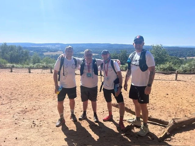 Four men standing outdoors on a dirt area with a fence and scenic landscape in the background. They are wearing casual hiking clothes, backpacks, and sunglasses.
