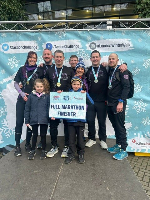 Group of eight people, including children, celebrating as full marathon finishers at a winter walk event with medals around their necks; they are standing in front of a blue backdrop with snowflake designs and event hashtags, holding a sign that read