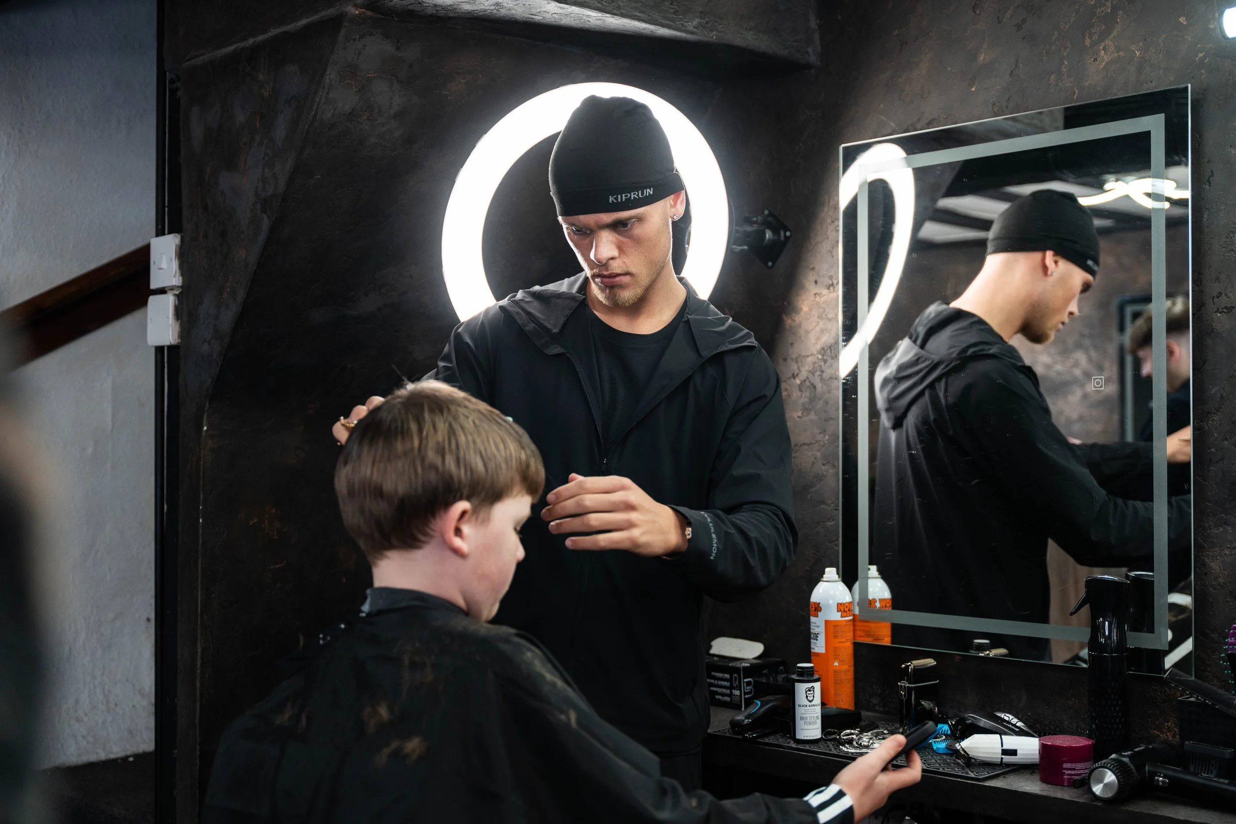 A young male barber in black clothing and a black beanie with 'KIPRUN' written on it cuts a young boy's hair in a modern barbershop with black walls, illuminated circular light, and a mirror reflecting the scene.