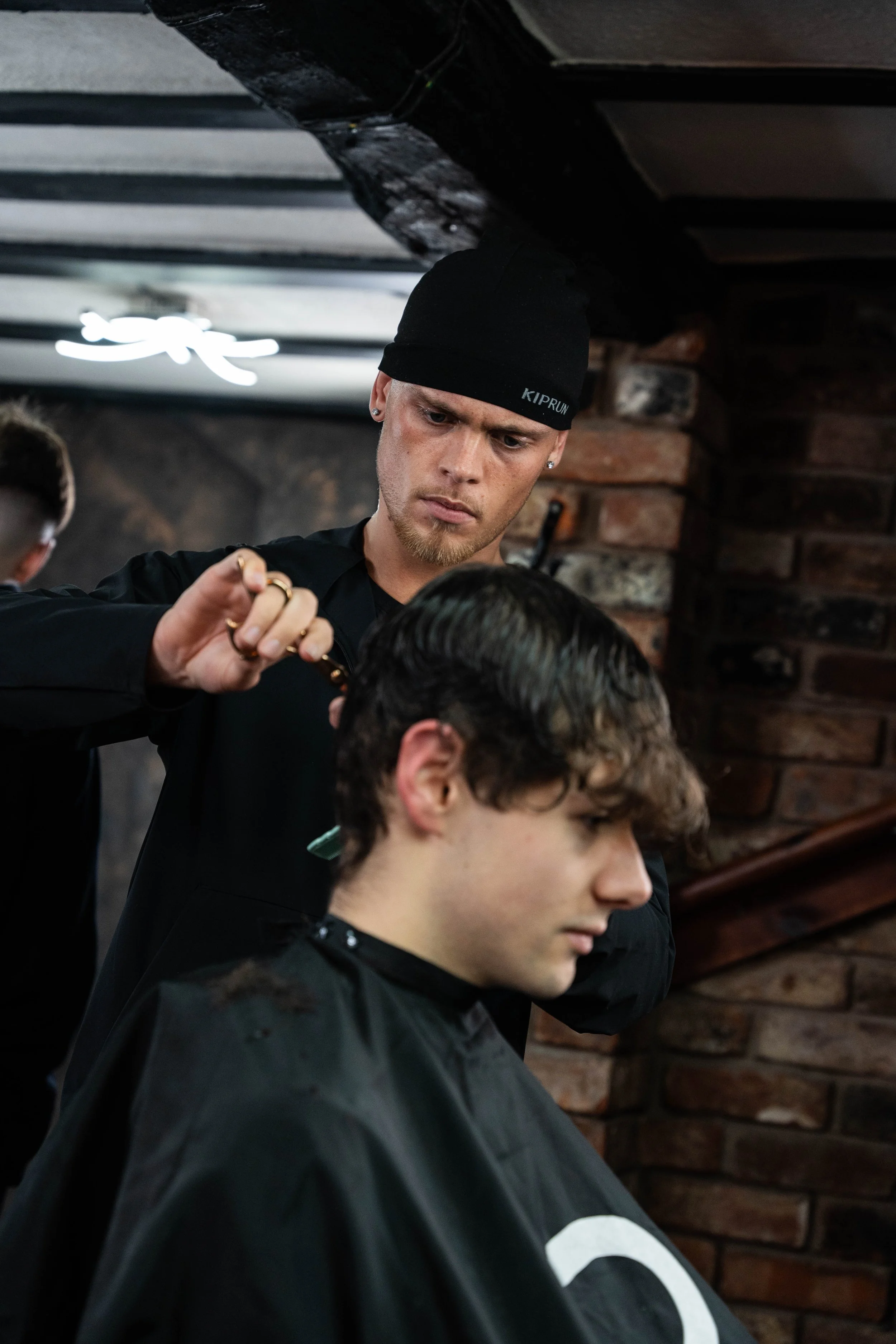 Barber cutting a young man's hair in a barbershop with brick walls and a ceiling fan.