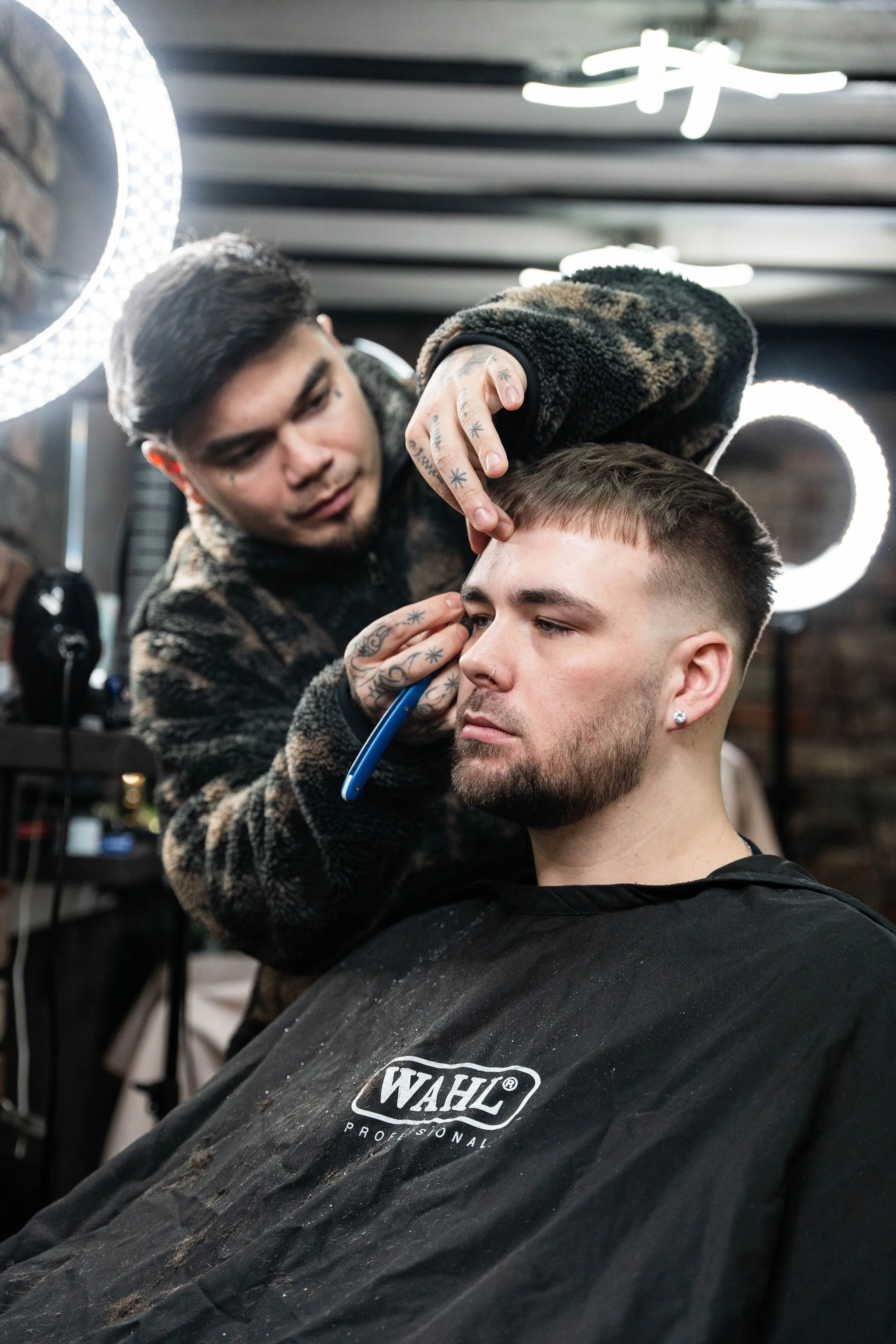 A barber with tattoos is cutting a man's hair in a modern barbershop, with ring lights and a brick wall in the background.