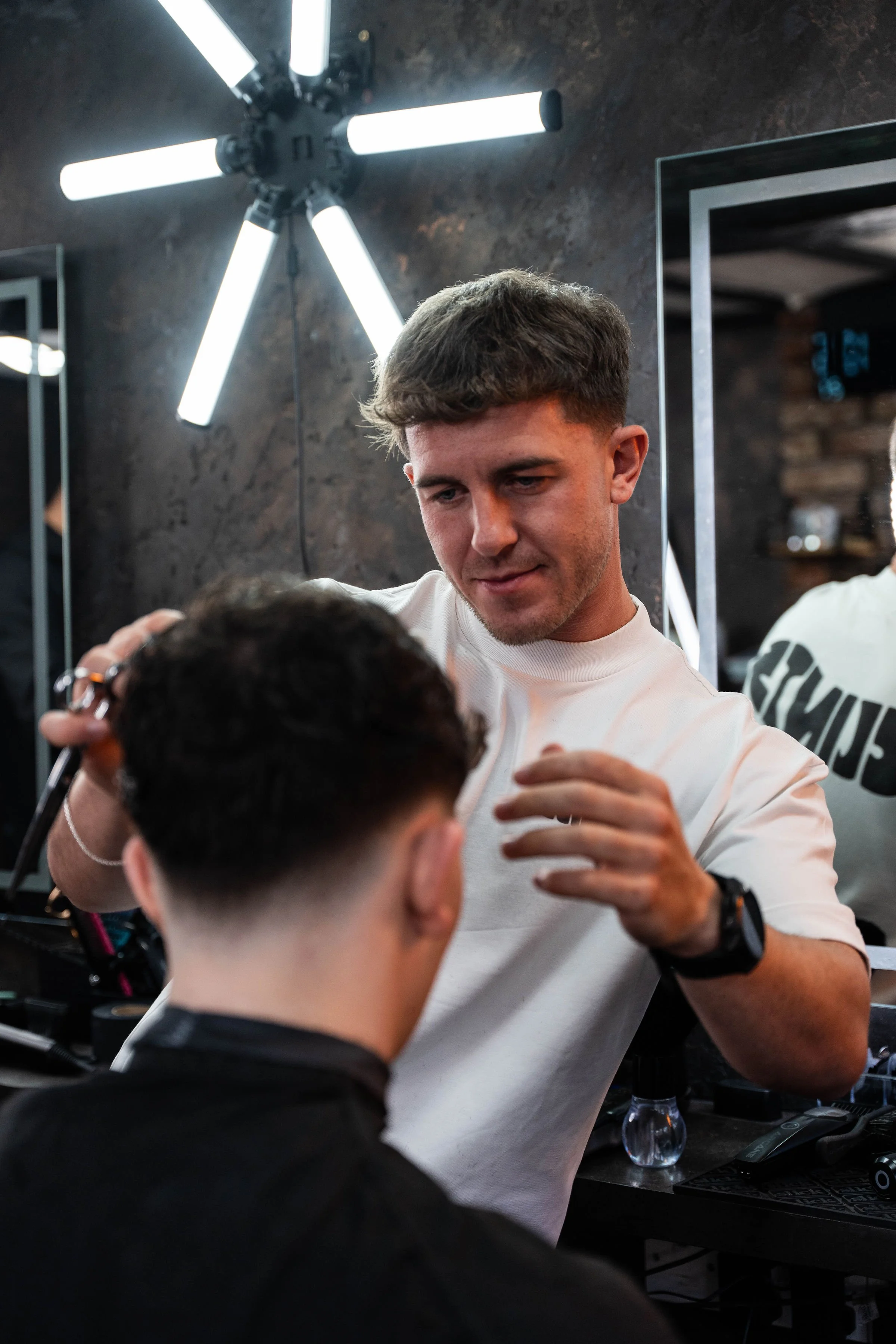 Male barber trimming a young man's hair in a modern barbershop with black walls and metallic accents.
