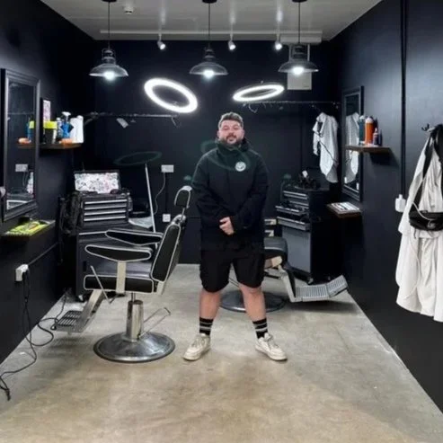 A man stands in a modern barbershop or salon with black walls and furniture, illuminated by circular and pendant ceiling lights, with barber tools and equipment around him.
