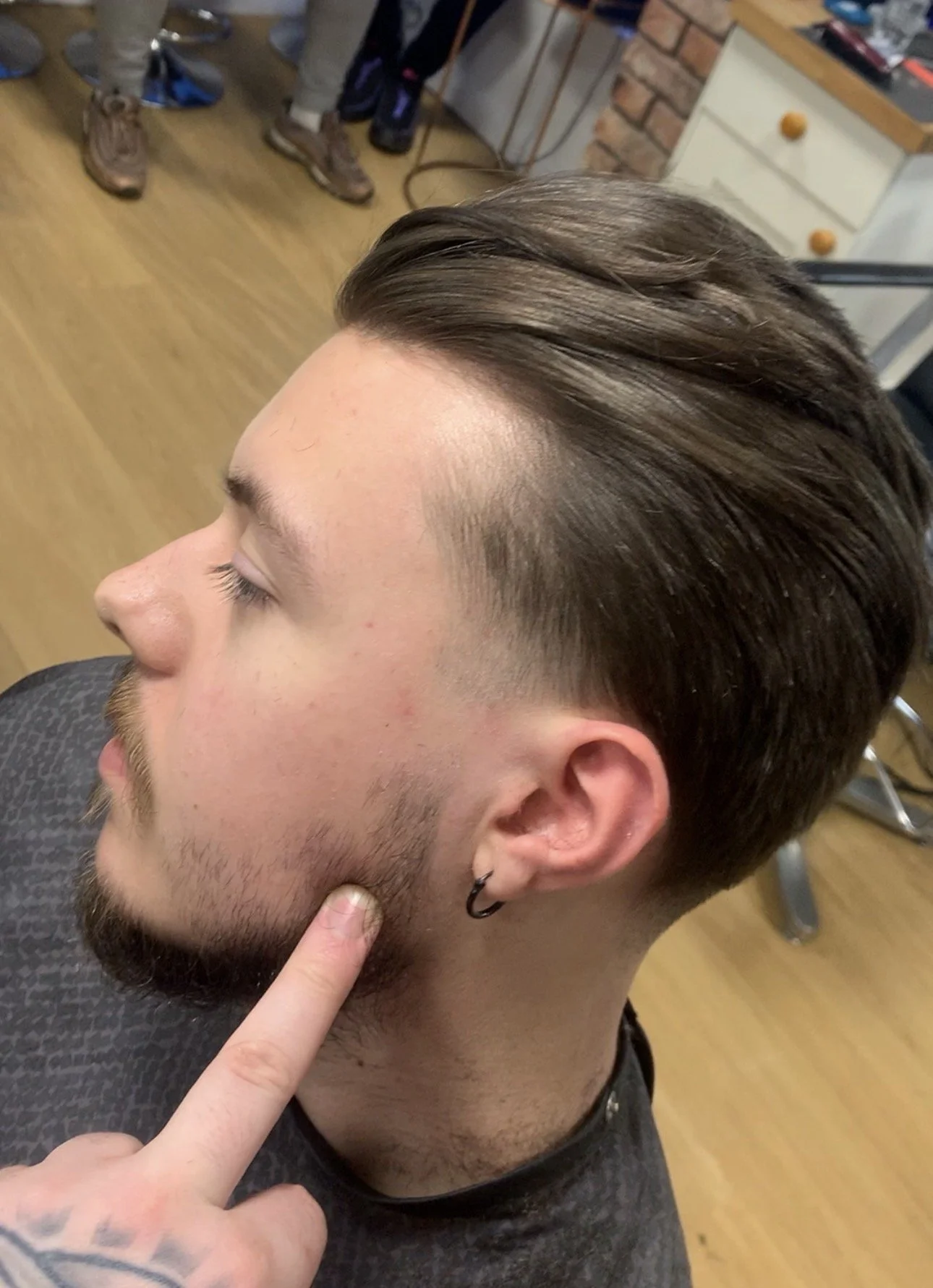A young man with styled brown hair and a beard is sitting in a barber's chair, getting a fade haircut. The barber's finger is touching his beard. The barber shop has a wooden floor, some footwear in the background, and a white dresser with orange kno