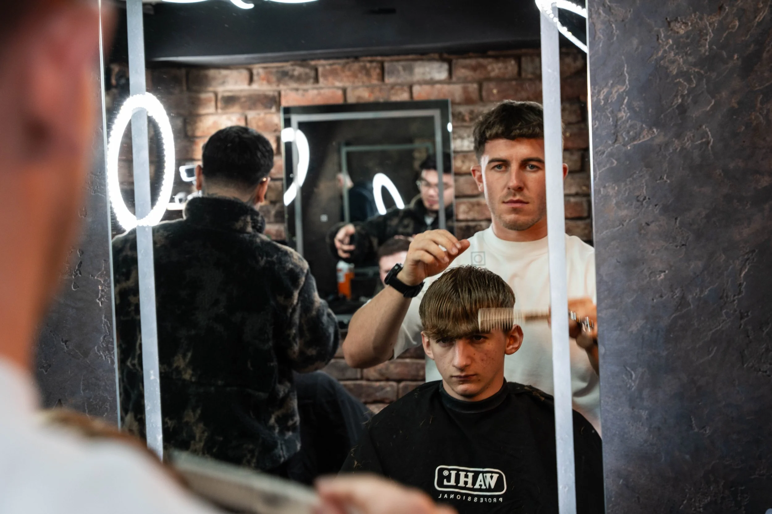 A young man gets a haircut at a barbershop. The barber, standing behind him, is styling his hair while looking into a mirror. The scene is reflected in a large mirror with a brick wall background, illuminated by a round ring light.