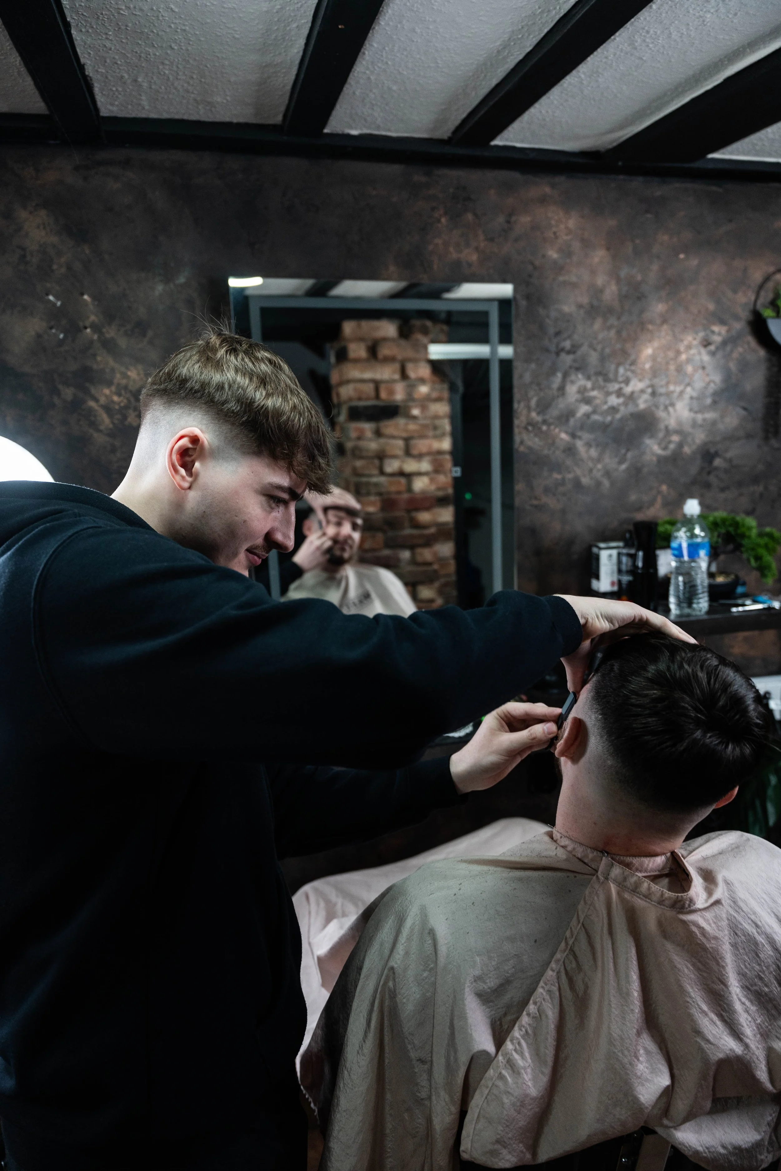 Barber giving a haircut to a customer in a barbershop with a brick wall and mirror behind them.