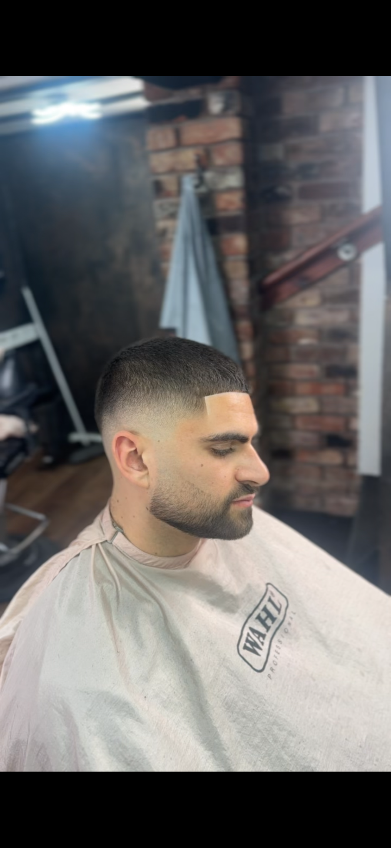A man with a fresh fade haircut sitting in a barbershop chair, wearing a barber cape with the logo WAHF, inside a barbershop with brick walls and barber tools in the background.