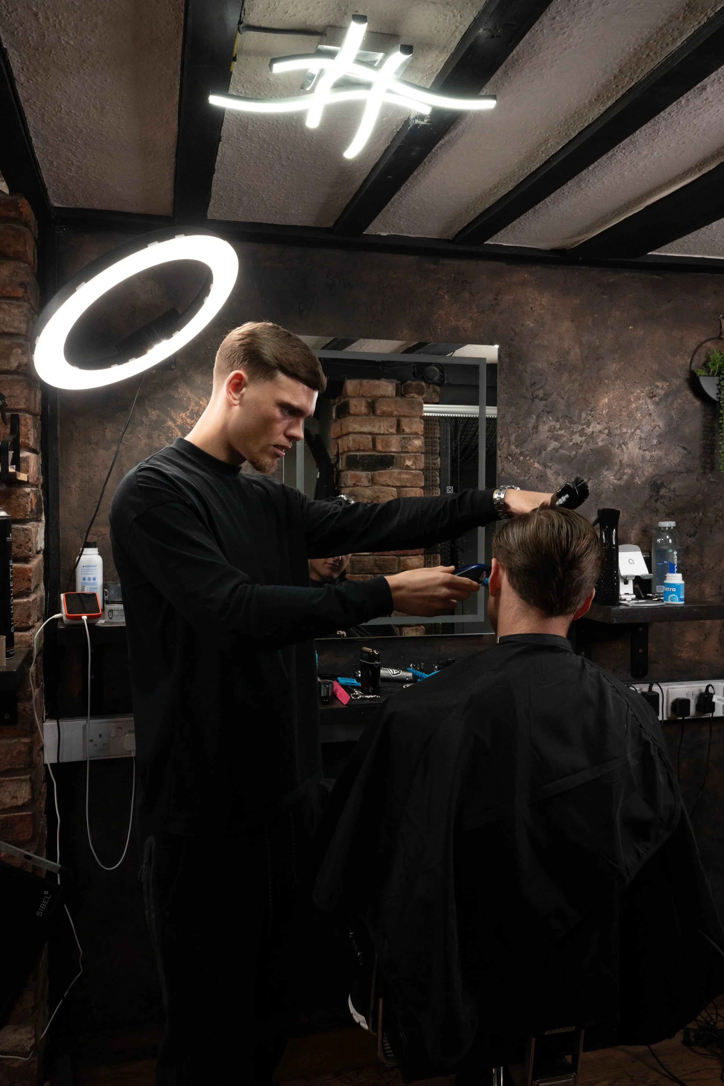 A barber is styling a customer's hair in a modern barber shop with brick walls and contemporary lighting.