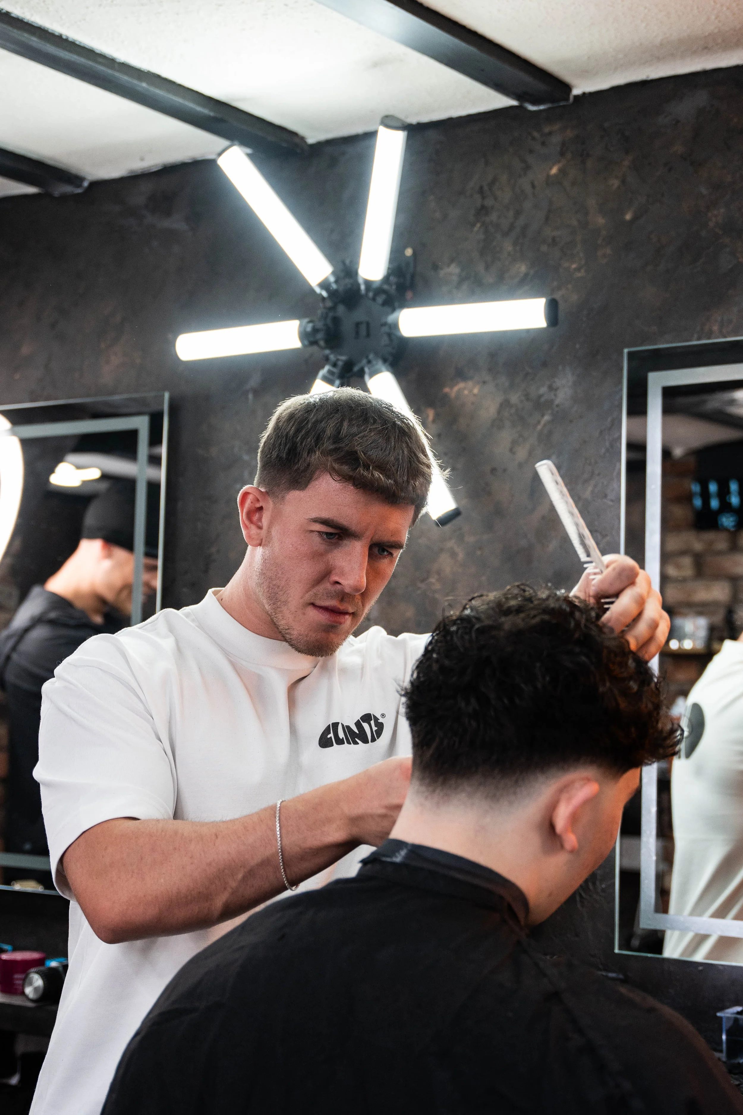 Barber cutting a man's hair in a modern barbershop with black walls and a unique ceiling light fixture.