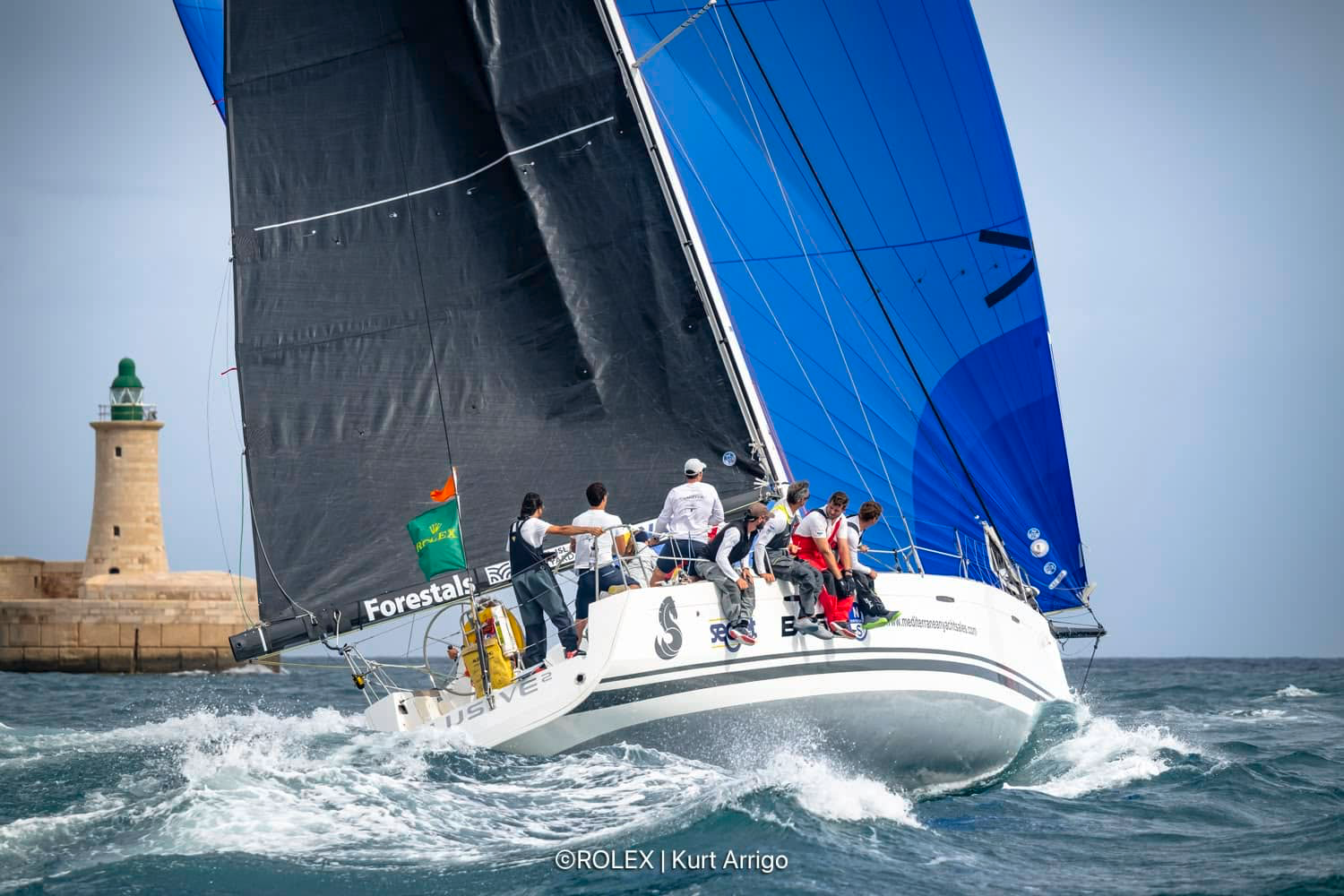 A sailboat racing on the water near a lighthouse, with a crew on the edge of the boat, some sitting and some standing, all handling the sails and equipment. The boat has black, blue, and white sails, and the background shows a clear sky and part of a stone lighthouse with a green top.