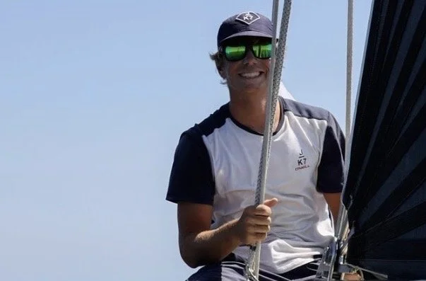 Man with sunglasses and cap smiling while on a boat, holding onto a rope.