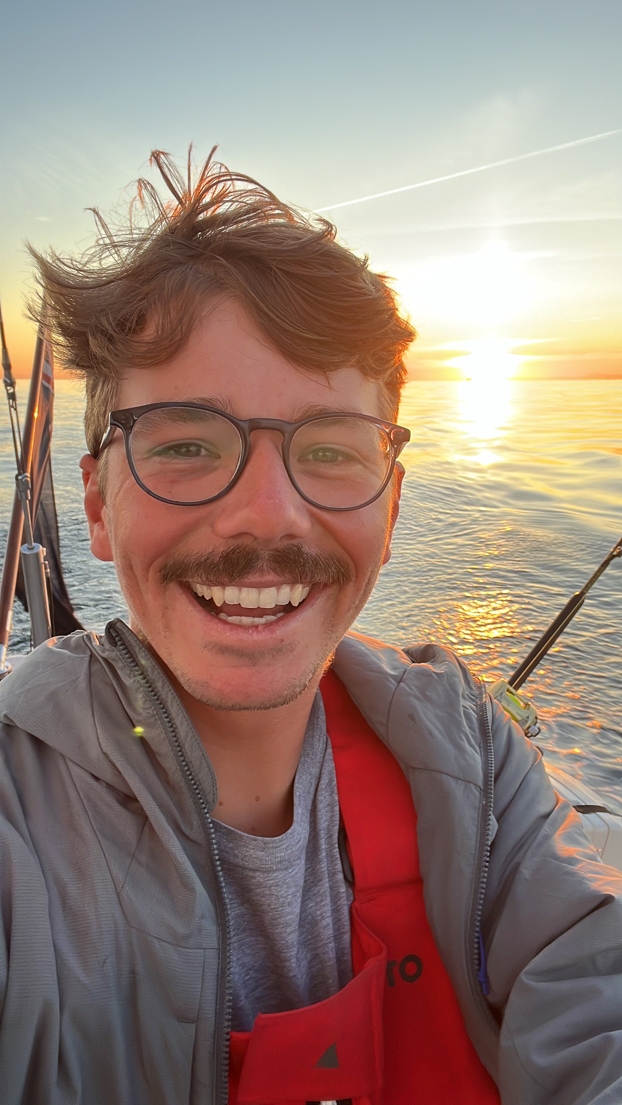 A smiling man with glasses takes a selfie on a boat at sunset, with water and the sun in the background.