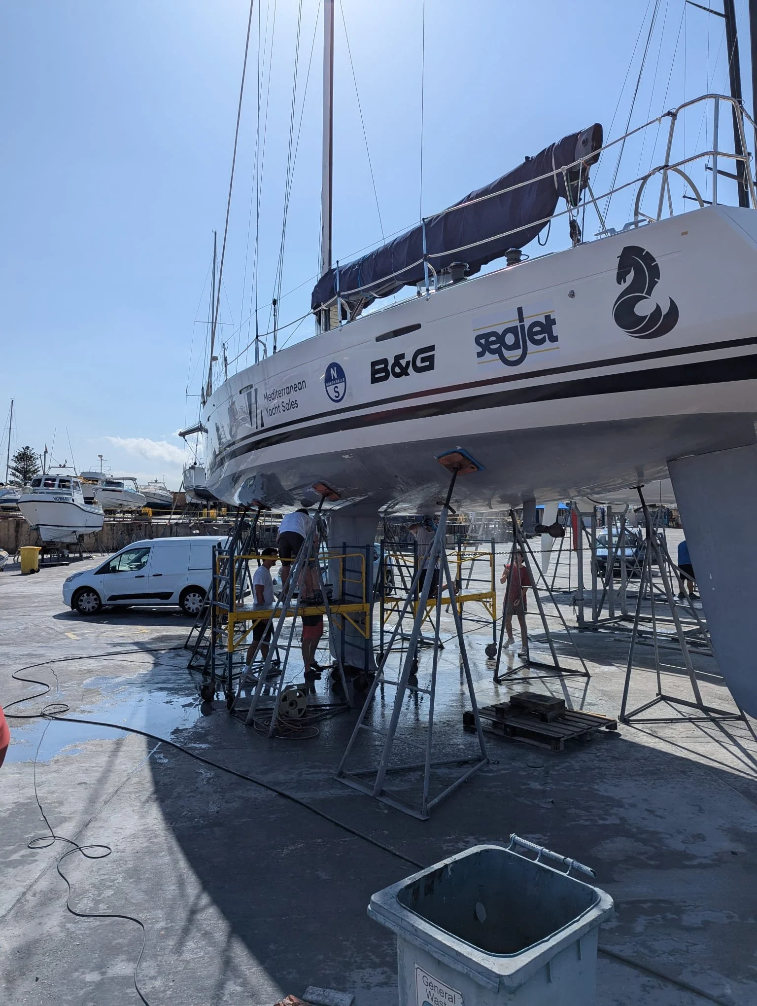 A large sailboat is out of the water at a boatyard, supported by stands for maintenance or repairs. Several workers are seen working around the boat, with some appearing to clean or inspect it. The sky is clear and sunny, and there are other boats and vehicles in the background.