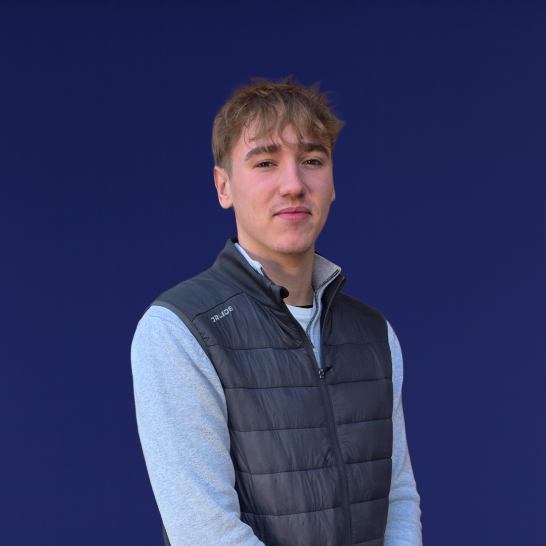 Young man with light brown hair wearing a gray long sleeve shirt and black puffer vest standing against a dark blue background.