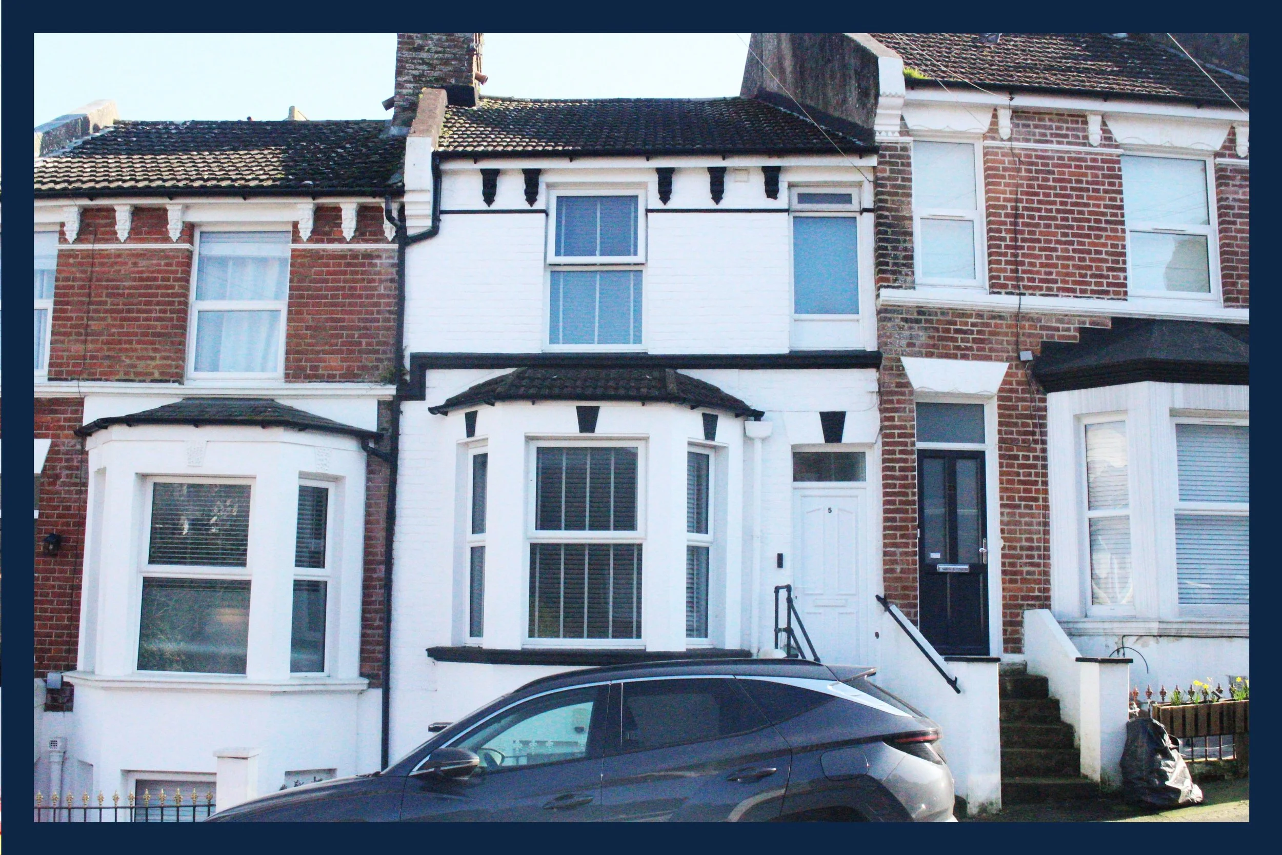 A row of brick and white painted houses with bay windows, steps leading to the entrances, and a parked gray car in front.