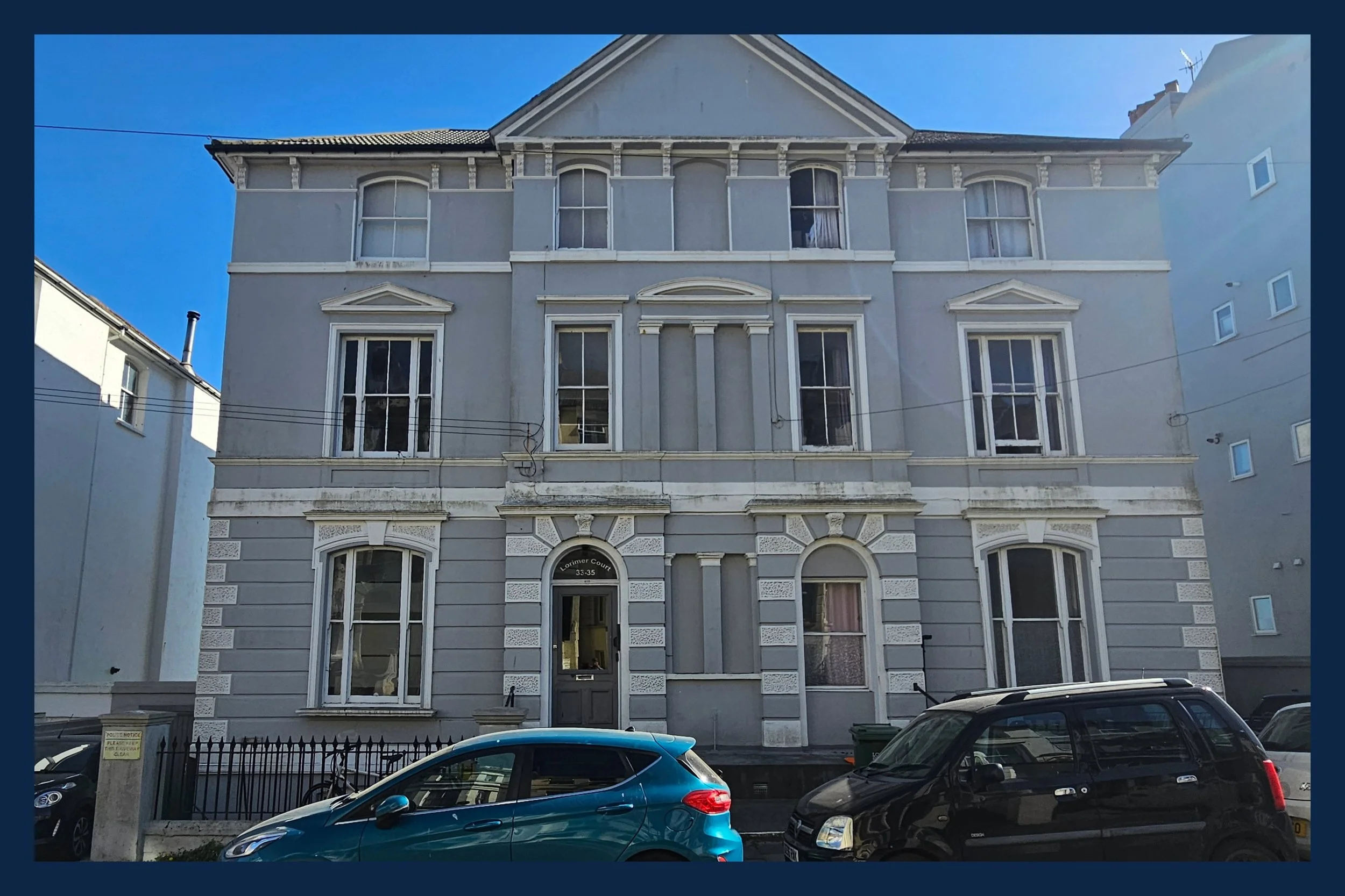 A four-story Victorian-style building painted in light blue with white decorative trim, featuring arched windows and ornate molding, situated in an urban setting with parked cars in the foreground.