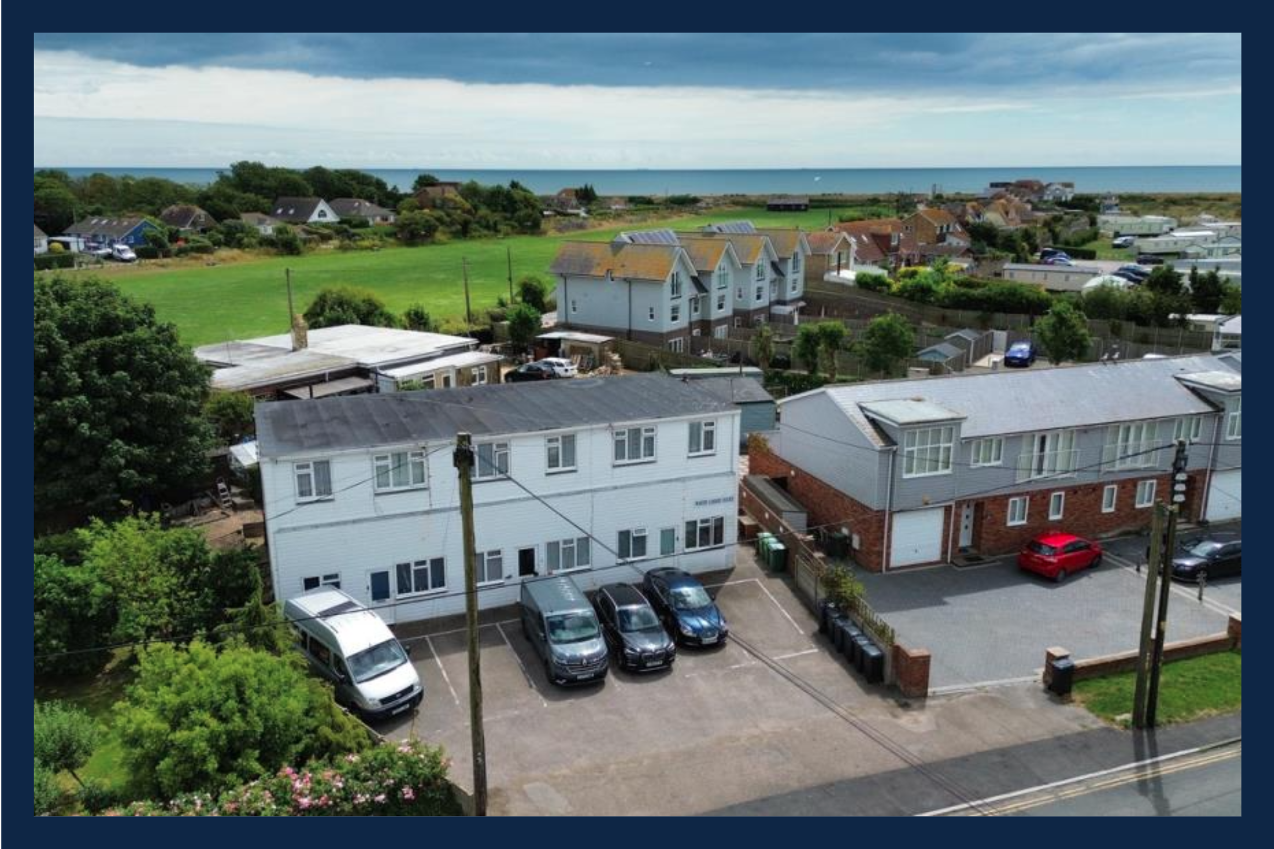 View of a residential neighborhood near the coast, showing two apartment buildings in the foreground, parking lots with cars, suburban houses, a green field, and the sea in the background under a cloudy sky.