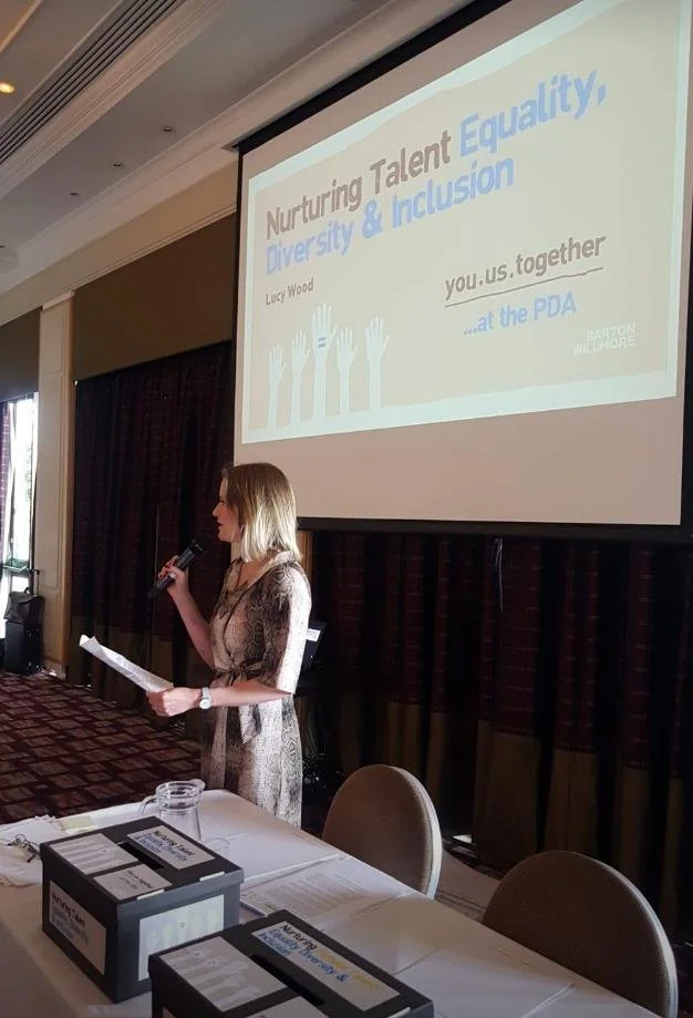 A woman giving a presentation at a conference, standing next to a large screen displaying a PowerPoint slide titled 'Nurturing Talent & Equality: Diversity & Inclusion'. She is holding a microphone and some notes. The room has curtains and tables wit