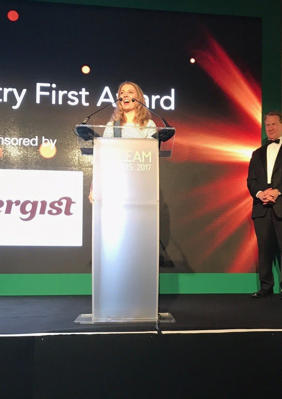 A photo of Rhiannon Smith speaking at a podium with a microphone during an award ceremony, with a man standing to the side on a stage with a large screen behind them displaying the text at the BREEAM awards. 