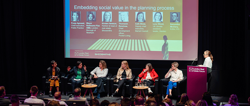 Panel discussion at the London Real Estate Forum with six women seated on stage and one woman standing at a podium, a large screen behind showing profile pictures and titles of speakers, audience members visible in foreground.